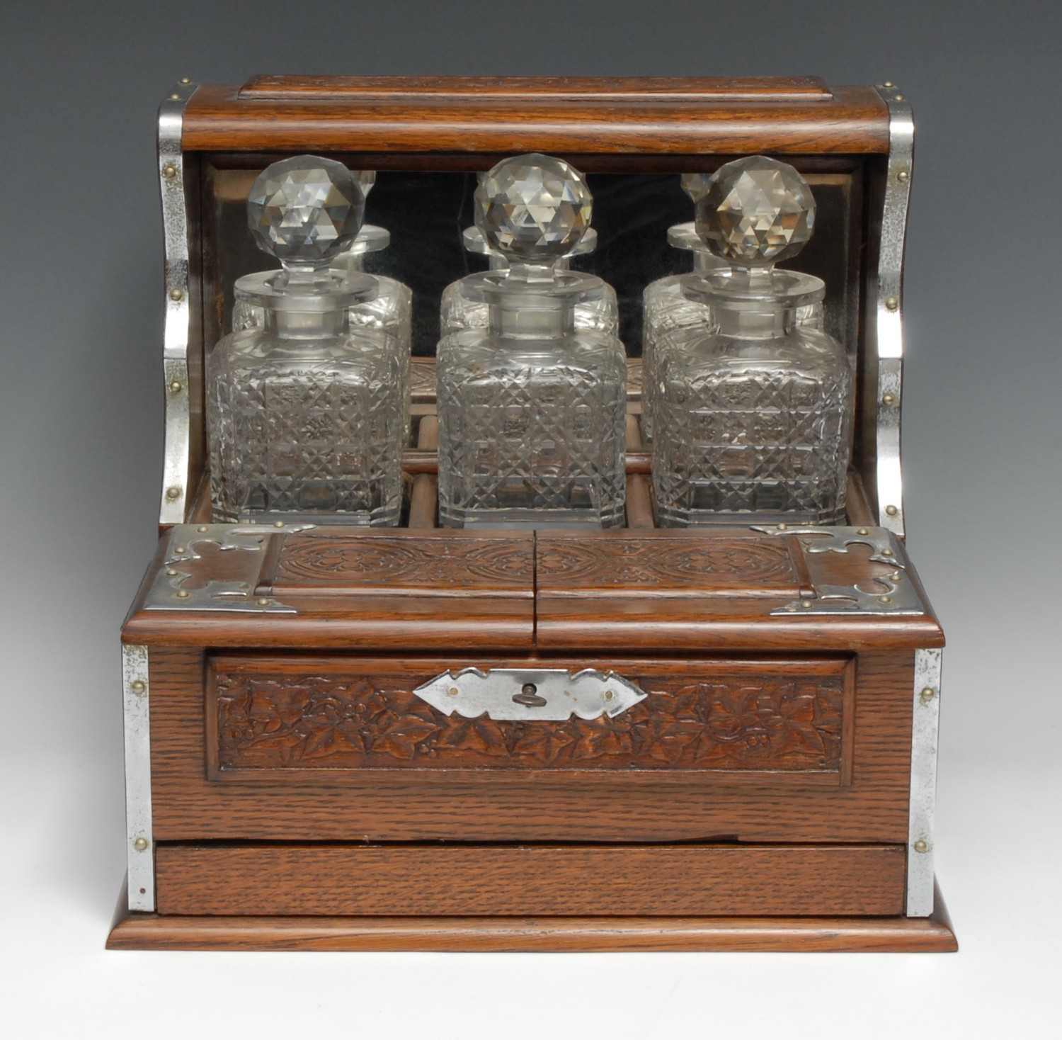 A late Victorian/Edwardian oak tantalus cabinet, the three hobnail decanters with prismatic stoppers, restrained within a mirrored niche, hinged twin covers enclosing drinking glass compartments, above a secret drawer fitted for games, carved with panels of fruiting vine, roundels and acanthus, carrying handles to sides, 33.5cm high, 35.5cm wide, c.1900