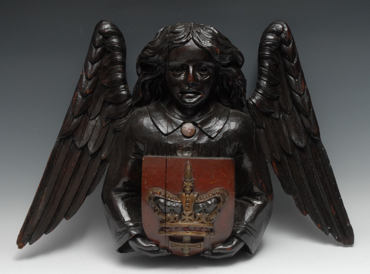 A 19th century polychrome painted softwood wall boss, carved half-length as a celestial figure supporting a shield, the emblazoned device a sword and Confessor's crown, 43cm high, 60cm wide