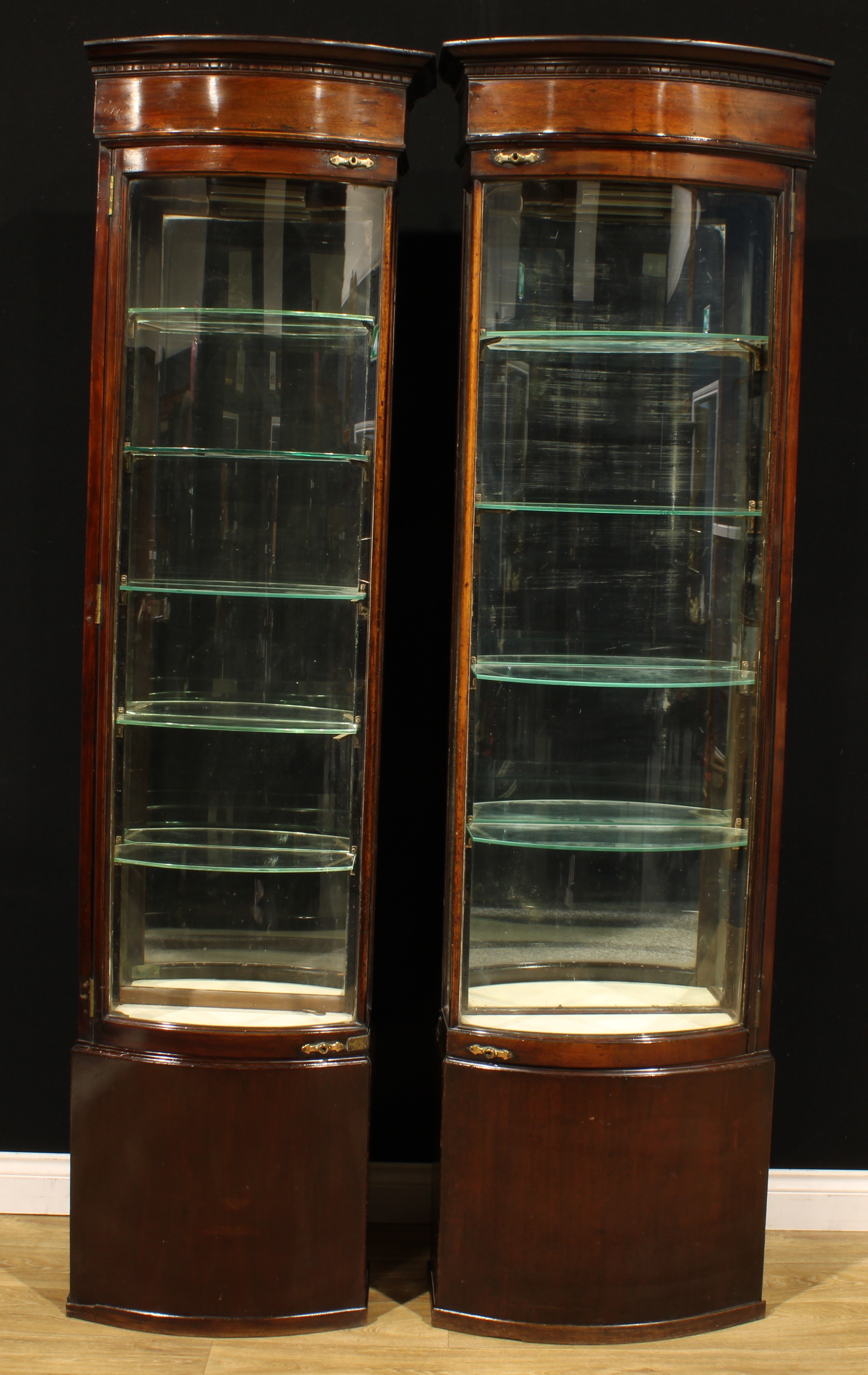 A pair of late Victorian mahogany bowfront shop display cabinets, each with outswept cornice with alternate flushed side, above a glazed door enclosing a mirrored back and shelves, three of the four locks stamped PATENT 3588, the largest 215cm high, 63cm wide, 30.5cm deep, the other 56.5cm wide, c.1895
