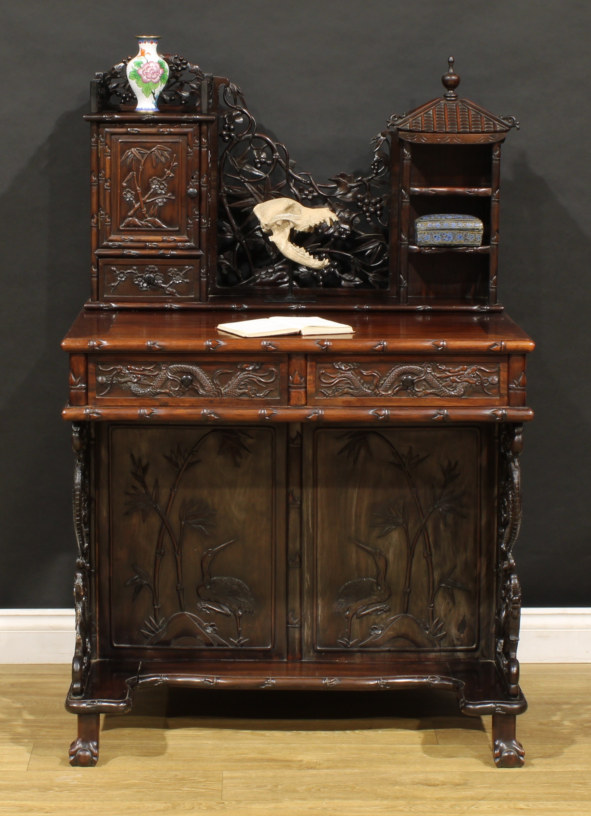 A Chinese export hardwood desk, shaped superstructure with panel door, small drawer and open shelves crested by a pagoda form pediment, centred by a shaped and pierced panel carved with leafy bamboo and fruiting vine, the projecting base with a pair of frieze drawers, each carved with a dragon, further drawers to sides, ball and claw feet, 131.5cm high, 86.5cm wide, 61cm deep