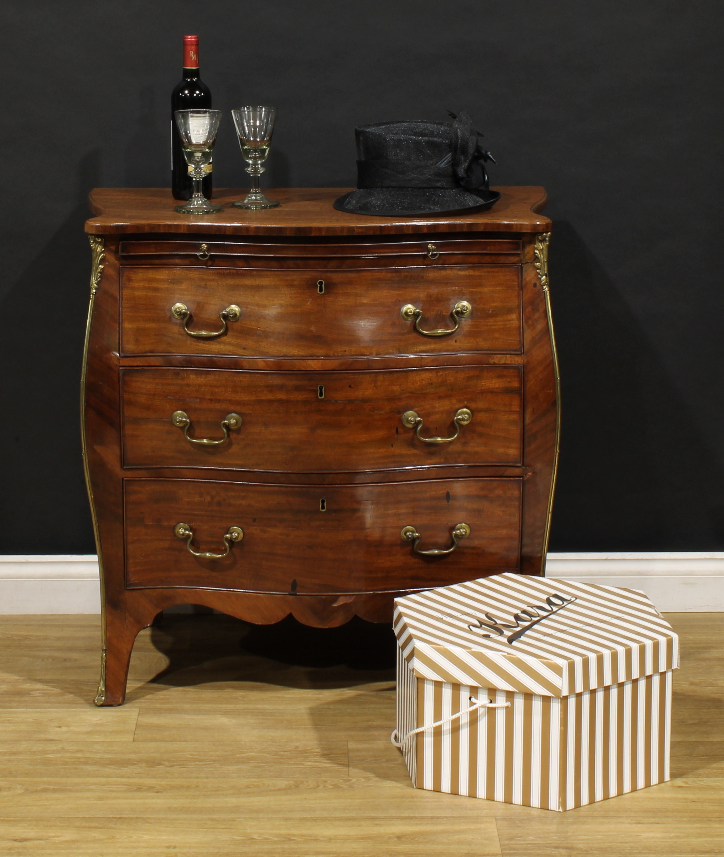 A 19th century gilt metal mounted mahogany serpentine bombe shaped bachelor’s chest, slightly oversailing top above a slide and three long cockbeaded drawers, brass swan neck handles, 87cm high, 80.5cm wide, 52.5cm deep