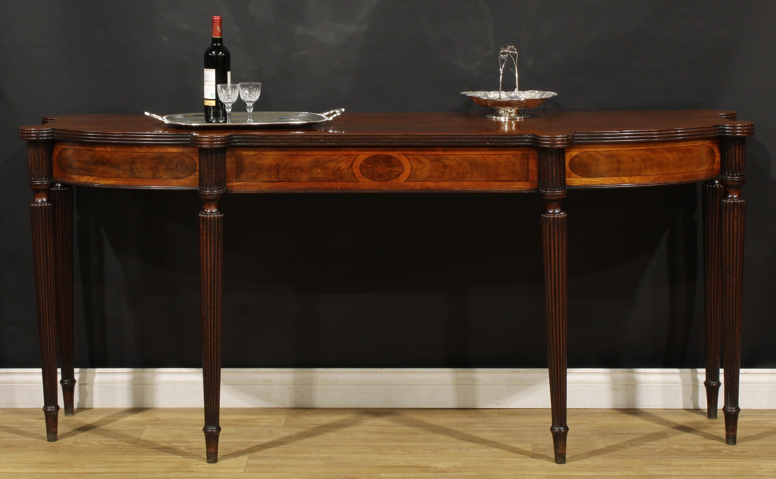 A George III mahogany serving table, in the manner of Gillows of Lancaster and London, slightly oversailing top with reeded edge above a satinwood and flame-veneered frieze with drawer to centre, fluted-capped tapered cylindrical reeded legs, 89.5cm high, 203cm wide, 69cm deep, c.1800
