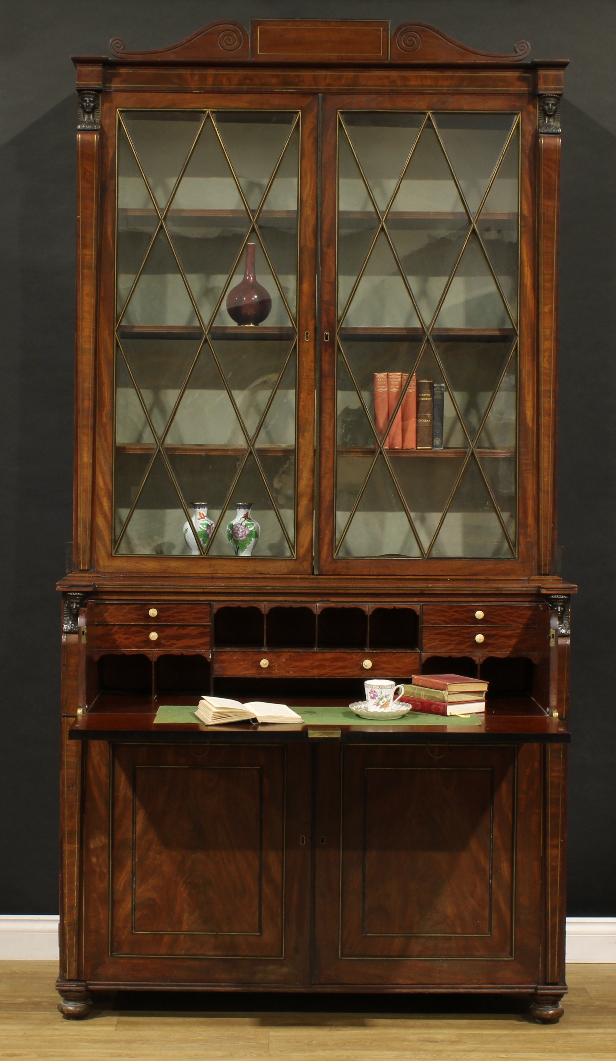 A Regency Egyptian Revival bronze mounted mahogany secretaire bookcase, outswept cornice with scroll pediment above a pair of astragal glazed doors enclosing three adjustable shelves, the projecting base with long fall front drawer enclosing a fitted interior, above a pair of panel doors enclosing three further long drawers, outlined throughout with brass stringing, turned feet, 244cm high, 123cm wide, 62cm deep, c.1820