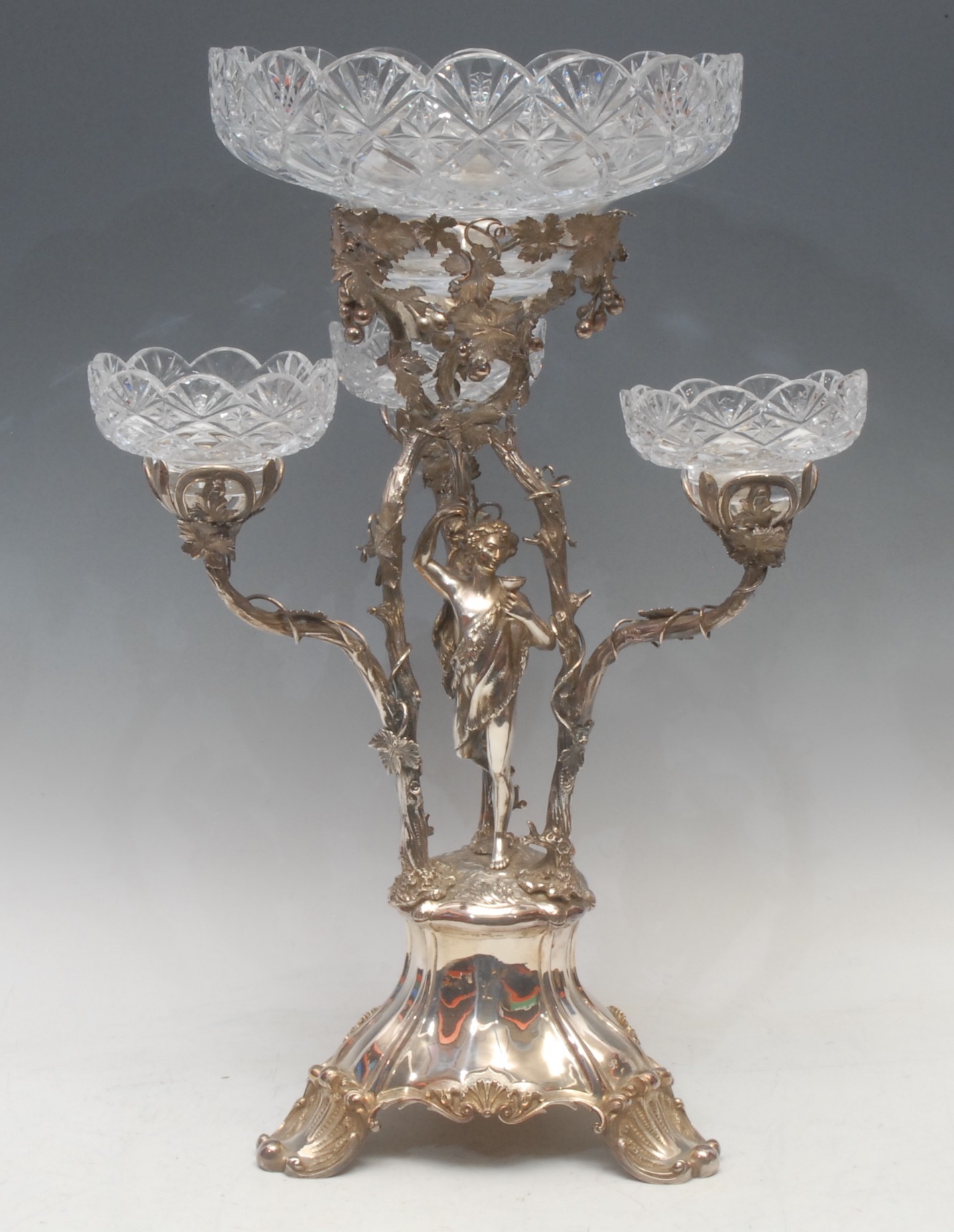 A Victorian silver three-branch table centrepiece or epergne, the central support cast with Dionysus, the god of wine, beneath a deep bower of fruiting vine, each branch naturalistically modelled and entwined with vine leaves, the trefoil base engraved with a stag crest, resting on three scalloped feet, fitted with four conforming, probably later, cut glass bowls, also three detachable sconces, 52.5cm, Smith, Nicholson & Co., London 1852, 2,050g