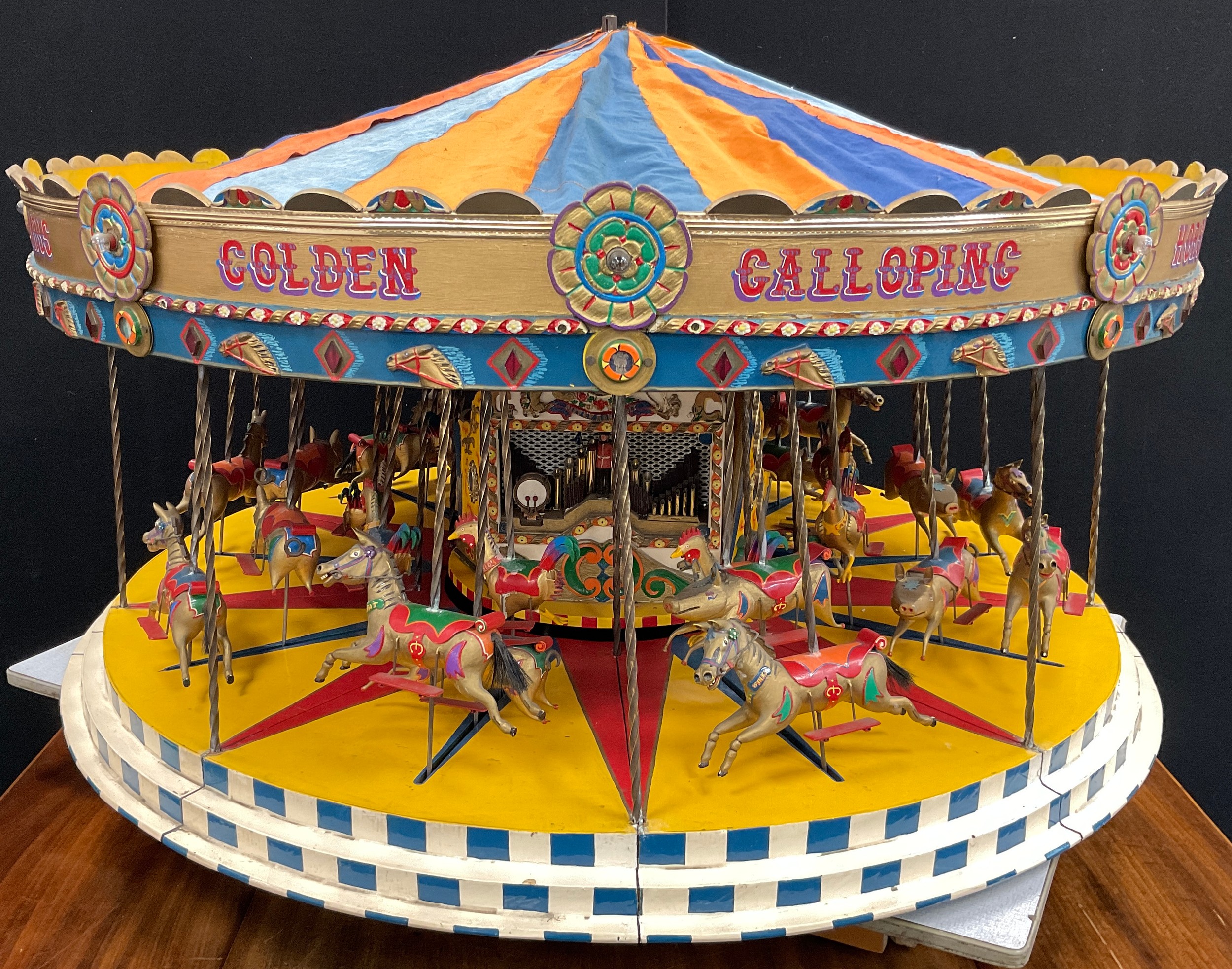 A handmade scratch built model, of a Fairground Carousel, made by Geoff Cocker from Eyam Derbyshire, over three years, the fully working model (needs rewiring) with horses, pigs, cockerels, painted in gold with multicoloured red, blue, yellow, green, orange and white detailing, the top with A Popular Pastime Famous Golden Galloping Horses, Cockerels and Pigs on Tour, approx 50cm high, 100cm diameter, with battery transformer (untested)