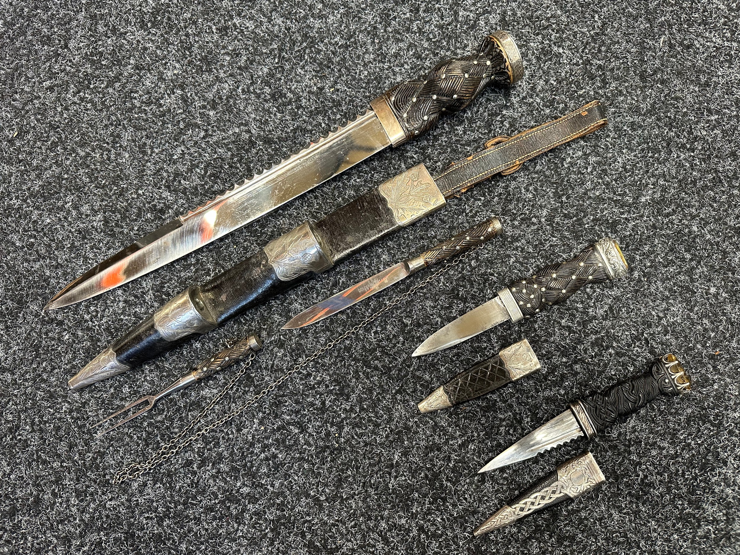 Scottish Dirk with 305mm long blade maker marked "Lockwood Brothers, Sheffield", carved wooden grip, white metal fittings. Overall length 420mm. Complete with scabbard with leather hanger. Engraved decoration of Thistles. Complete with Knife and Fork secured with a chain, matching makers mark. Along with two Sgian Dubh knives, one with matching design from the same maker, with 80mm long blade, overall length 176mm. And another no maker mark with 83mm long blade, overall length 170mm with silver fittings Hall marked for Edinburgh 1937. (3)