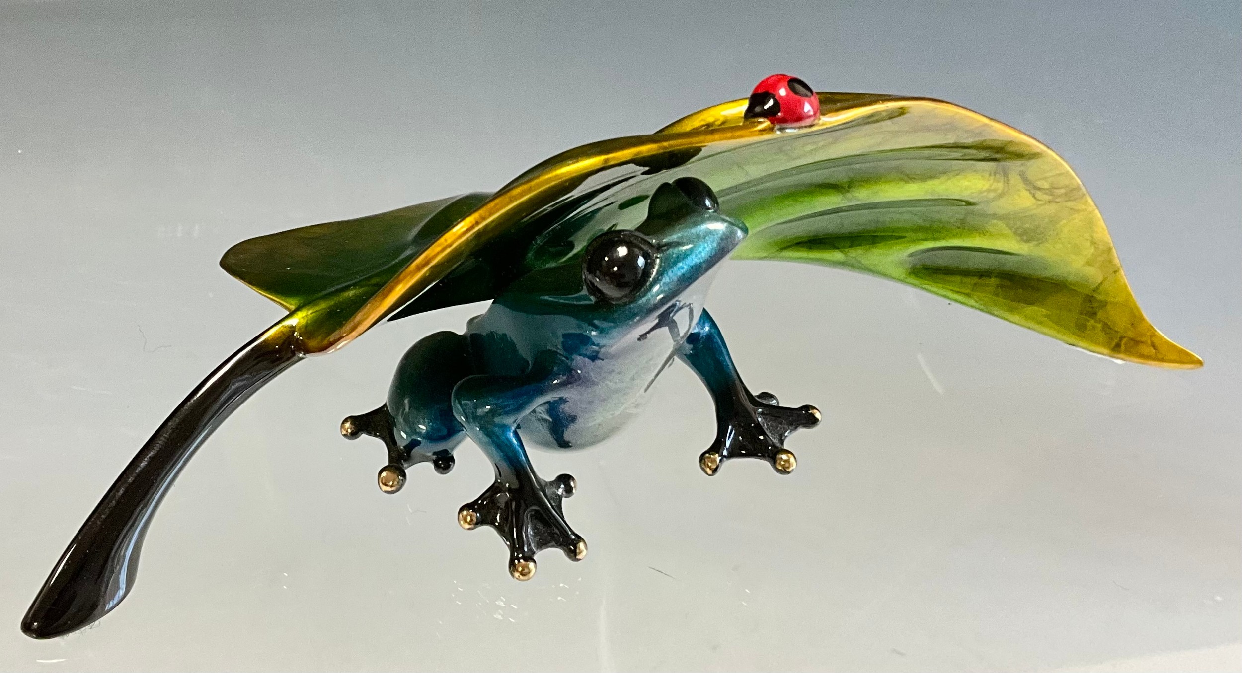 Tim 'Frogman' Cotterill; a limited edition lost wax cast bronze model 'Under Cover', of a frog with a blue hue patina, resting under a golden green leaf, a Ladybird to the edge, 718/2000, signed, numbered and impressed on the underside, approx 6cm high, 17cm wide, with certificate Mulberry Hall, 17/10/11.